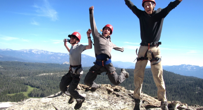 Three students wearing helmets and harnesses leap into the air in celebration. They appear to be on a summit, high above a wooded landscape. There are mountains in the background.